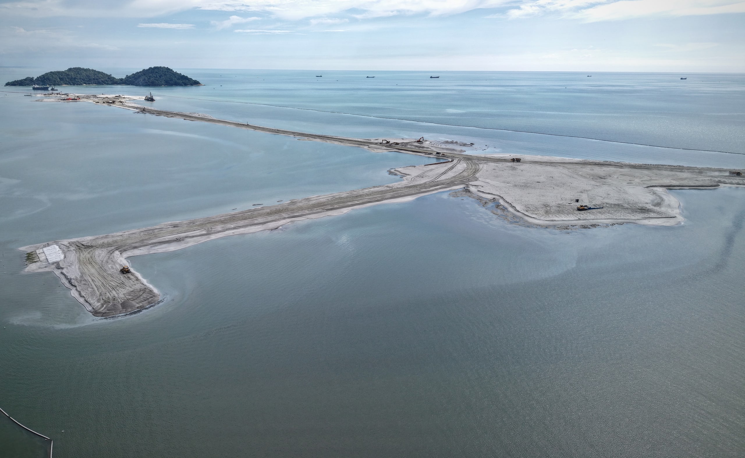 Aerial view of the Silicon Island reclamation: a T-shaped sand platform in the sea, with Pulau Rimau on the horizon